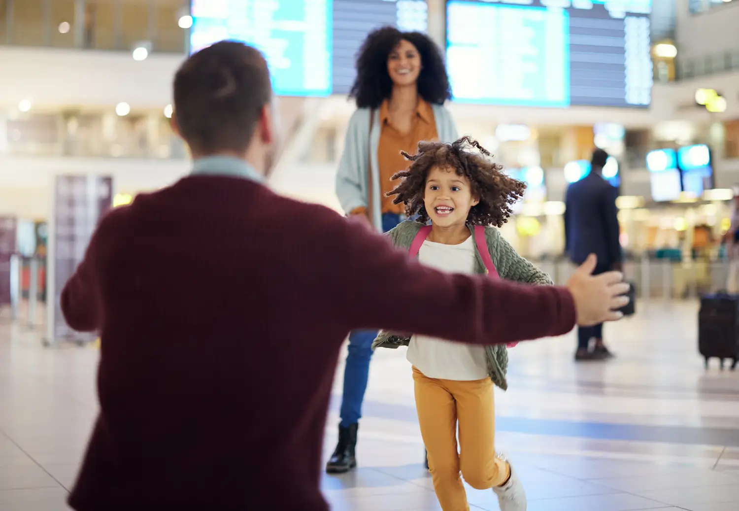 A young child with curly hair is joyfully running towards a person (whose back is to the viewer) with open arms for a hug, in an airport terminal. Another woman, possibly the mother, stands smiling in the background. The scene captures a heartwarming family reunion or greeting, with airport screens and other travelers in the blurred background.